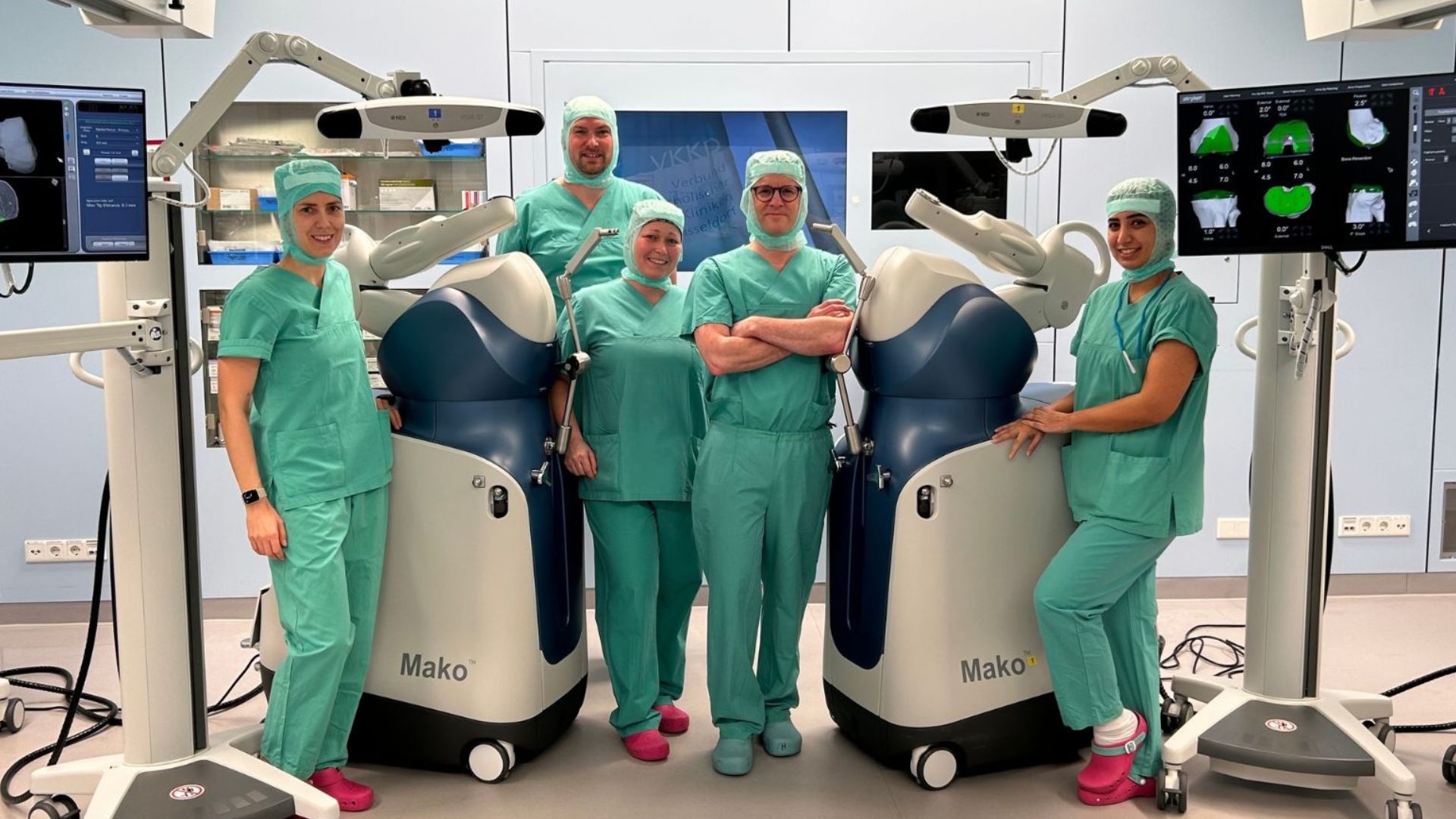 Fünf Mitglieder des Teams der Orthopädie stehen in grüner OP-Kleidung vor den OP-Robotern im OP-Saal.